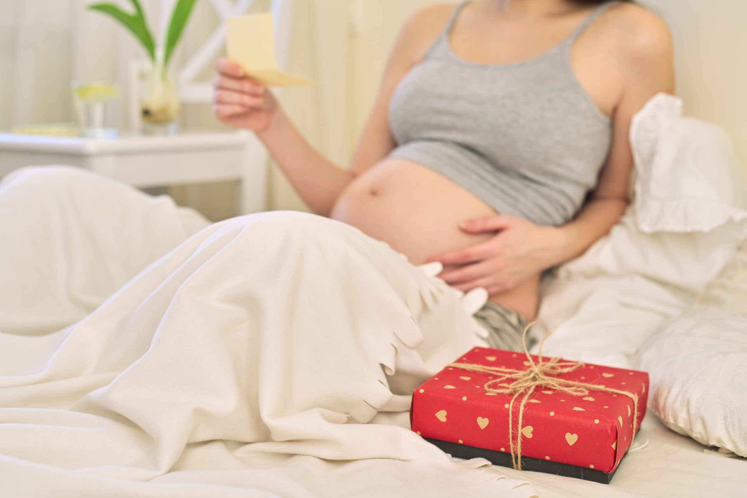 young-beautiful-pregnant-woman-with-gift-box-female-sitting-home-bed-reading-greeting-card (1)